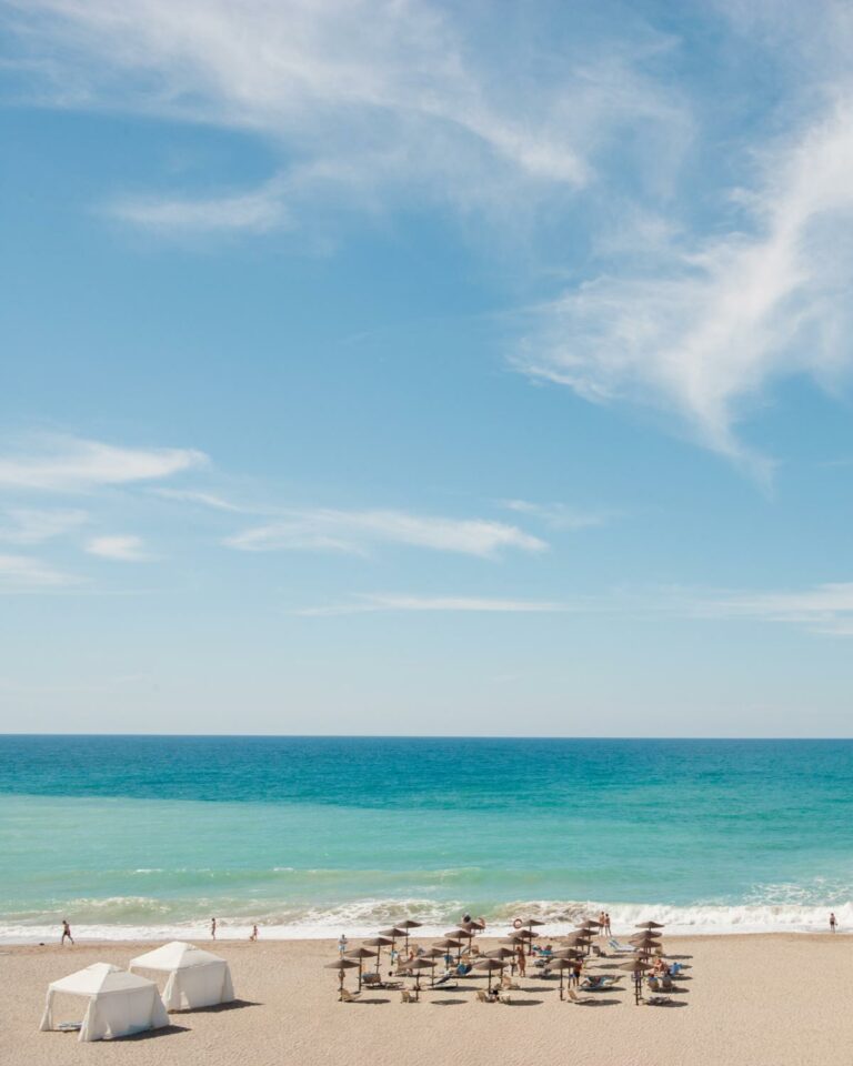 Wide view of a sandy beach with sun loungers, umbrellas, white cabanas, and turquoise ocean under a blue sky with wispy clouds.