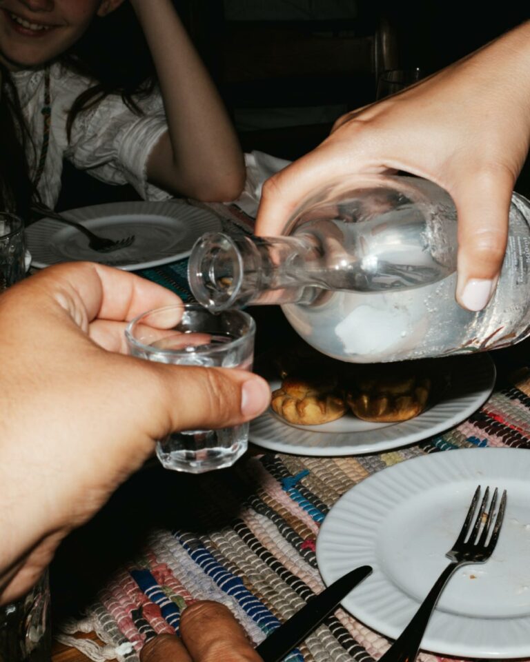 Hands pouring raki from a glass bottle into a small drinking glass beside a plate of food.