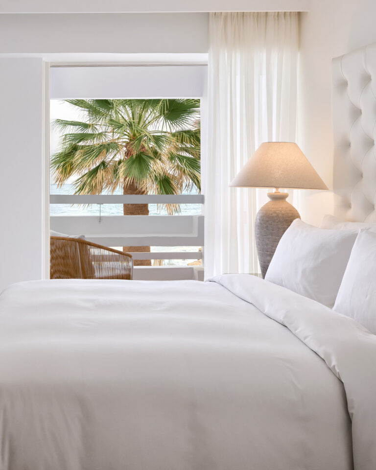 Grecotel LUXME White Crete White Family Suite bedroom with sea view, palm tree and bedside lamp.