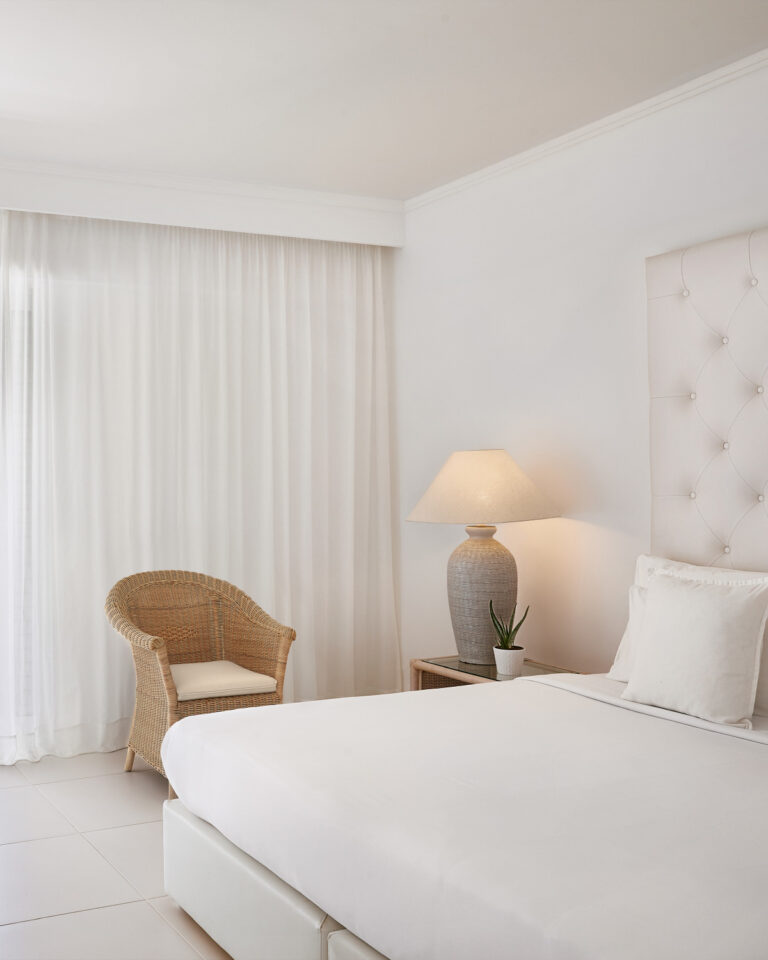 Grecotel LUXME White Crete Family Apartment bedroom with king bed, wicker chair and bedside lamp.