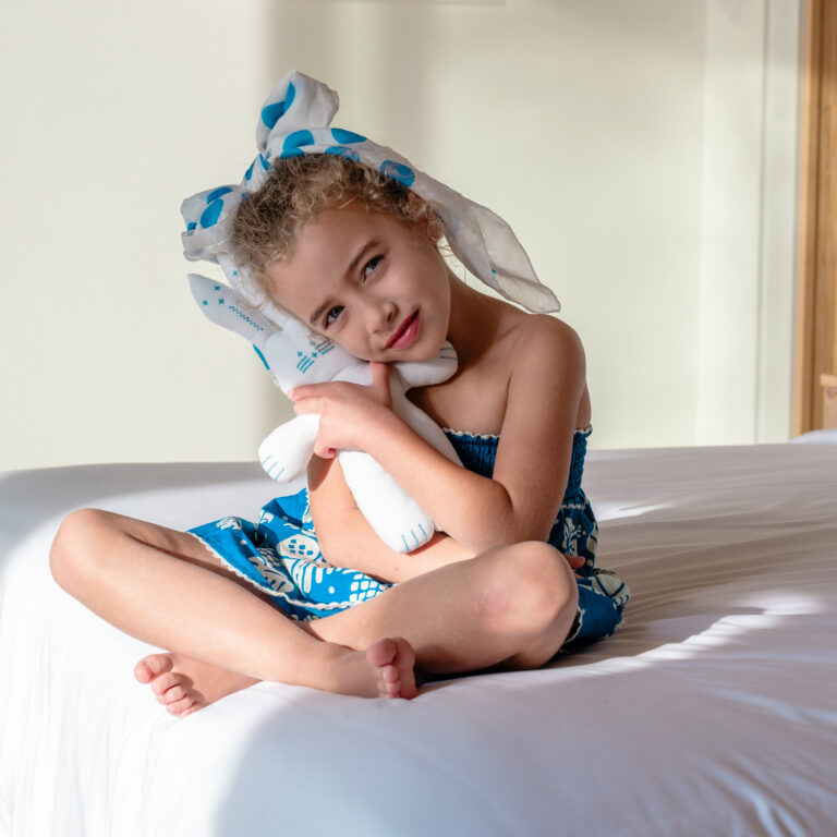 Child sitting on a bed with a soft toy in a Grecotel LUXME White Crete Yali Seafront Suite.