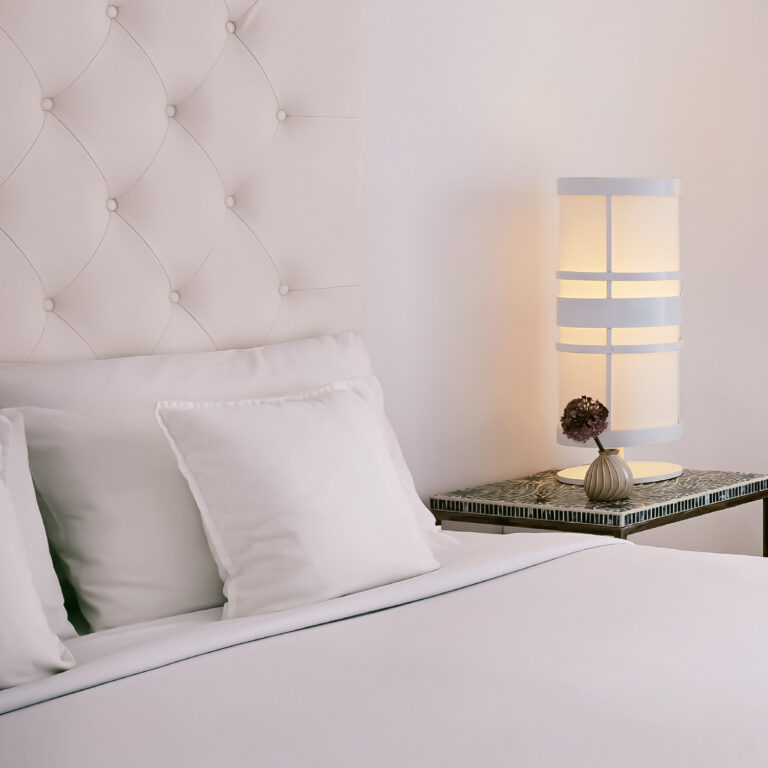 Grecotel LUXME White Crete swim-up guestroom bedroom with white bed and bedside lamp.