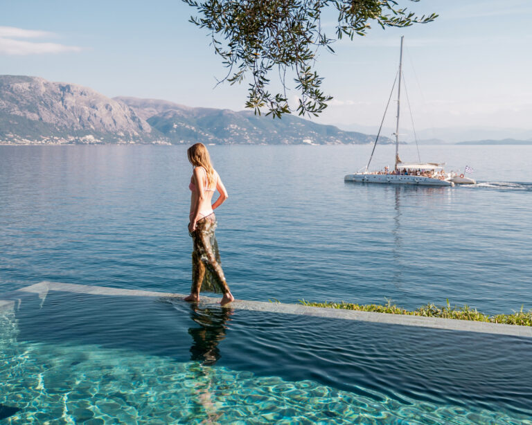 Woman walking beside an infinity pool overlooking the Ionian Sea with mountains and a sailboat in the background.
